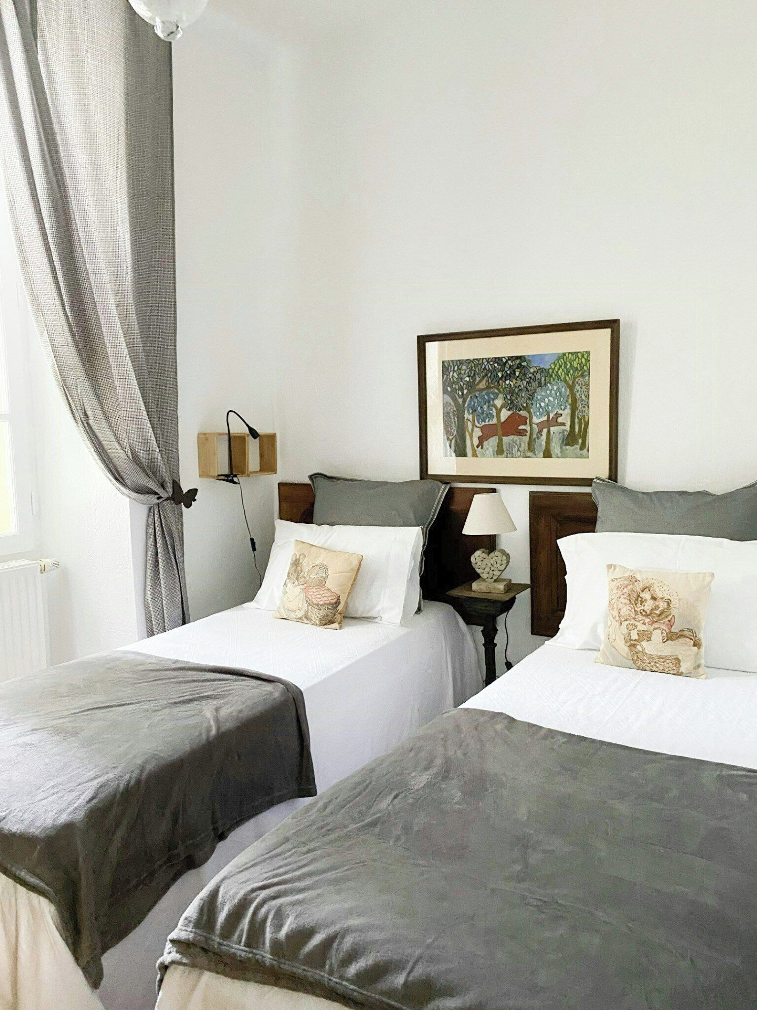 Une chambre d’hôtel avec deux lits, une table de chevet, une lampe, un tableau encadré et une fenêtre avec des rideaux.