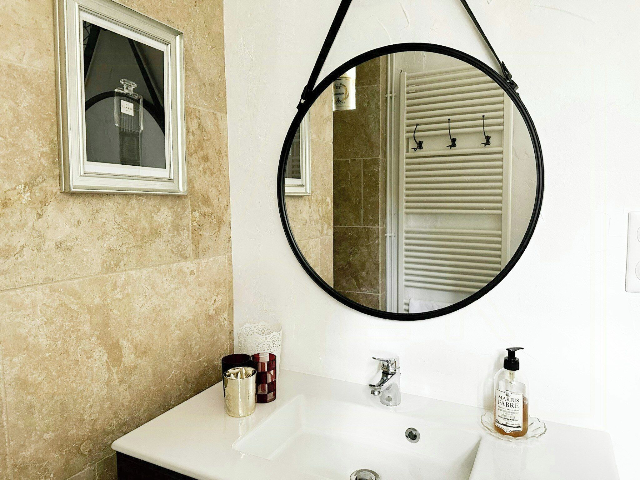 Un baño con un espejo redondo, una botella de Chanel con estuche, un lavabo con grifo y una botella de jabón Marius Fabre.