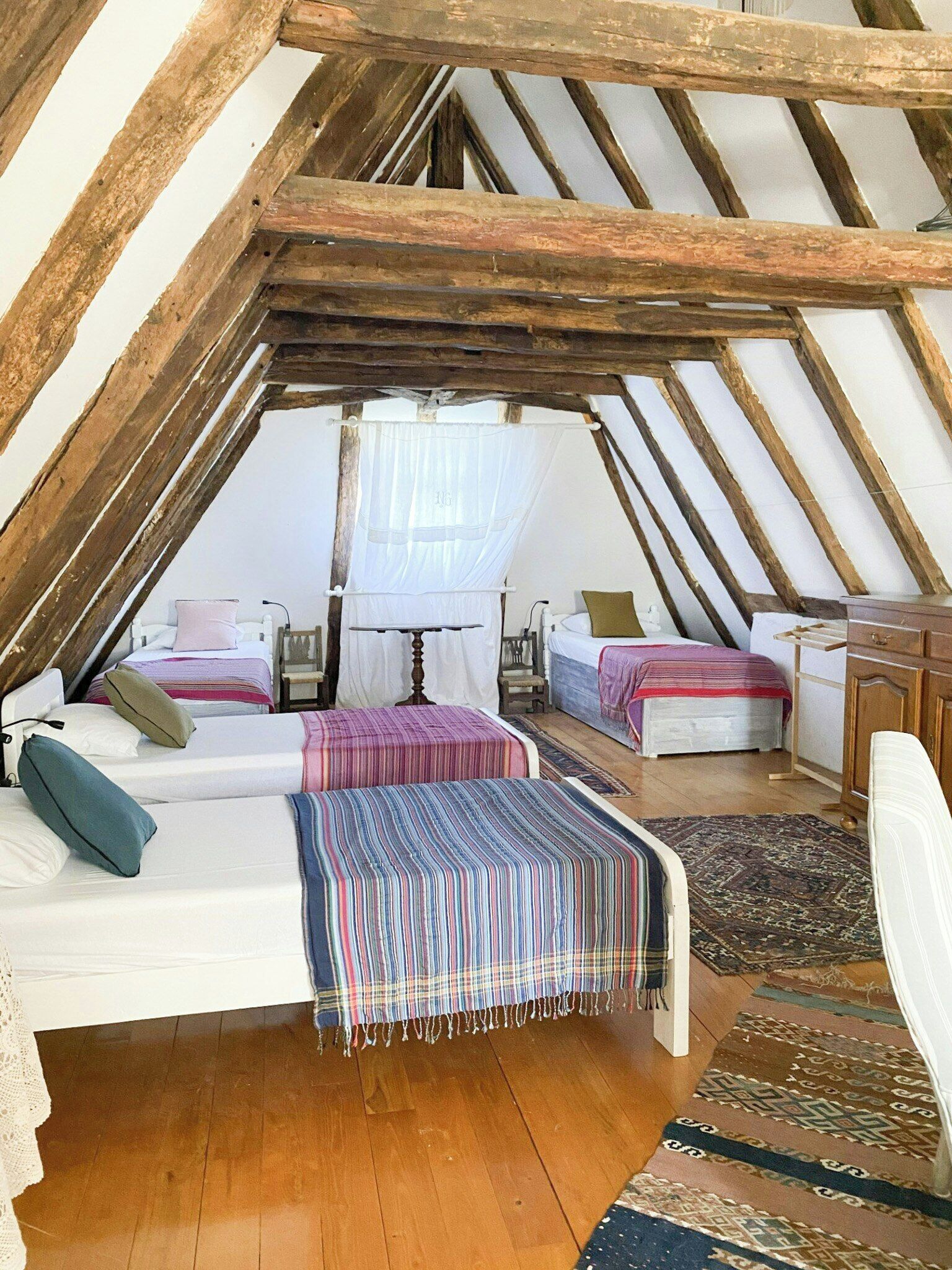 Une chambre confortable située sous les combles, avec des poutres apparentes, deux lits, une table de chevet et une grande fenêtre ornée d’un rideau léger.