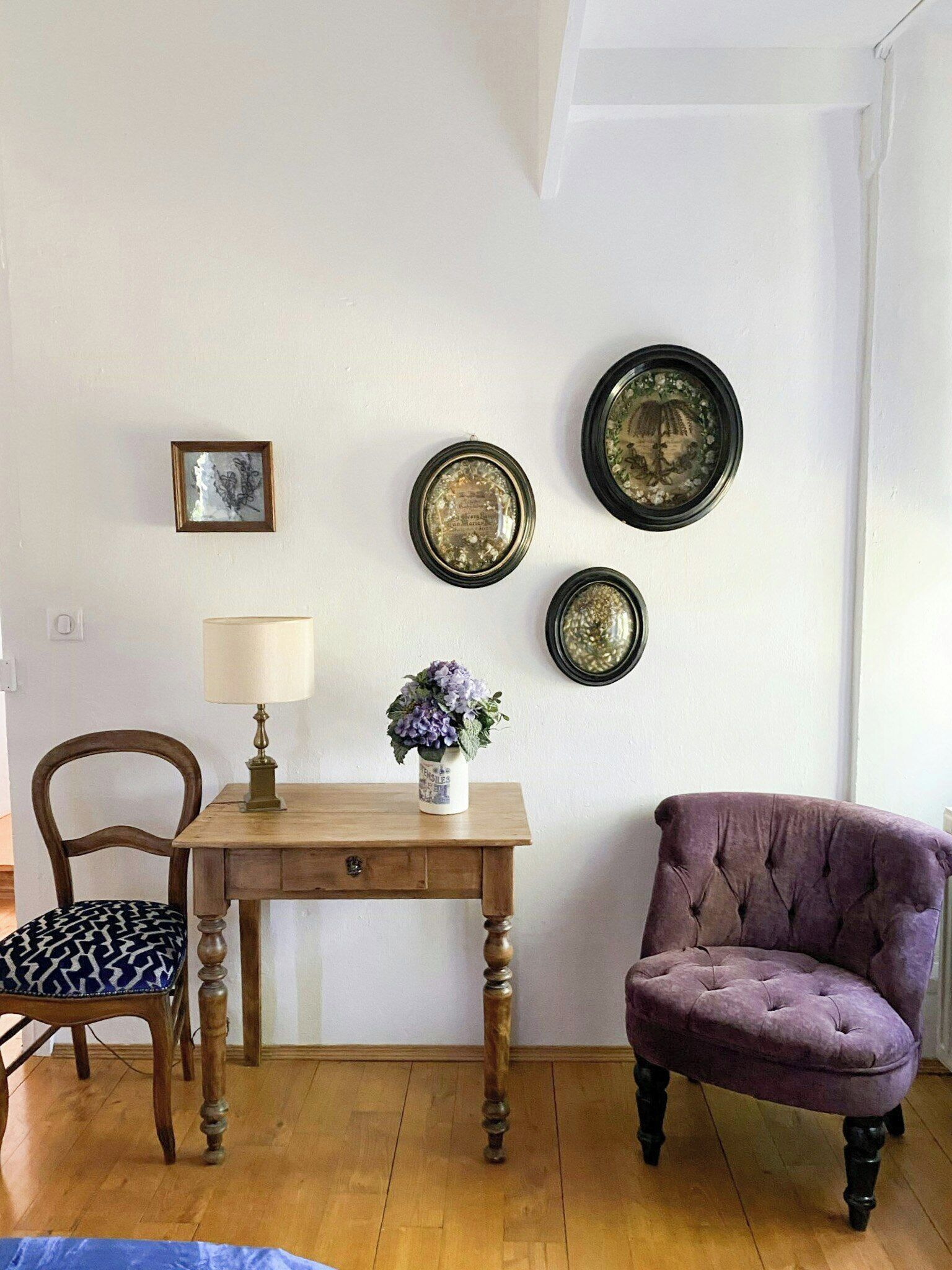 Una habitación con una mesa de madera, un sillón morado, una lámpara, un jarrón con flores y tres placas decorativas colgadas en la pared.