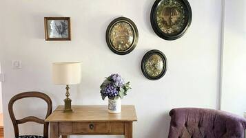 Una habitación con una mesa de madera, un sillón morado, una lámpara, un jarrón con flores y tres placas decorativas colgadas en la pared.