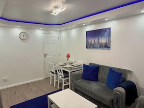 Ein modernes Wohnzimmer mit grauem Sofa, blauen Kissen, einem Esstisch mit Stühlen, einer Uhr und einem Wandbild mit einer Sternennacht.
