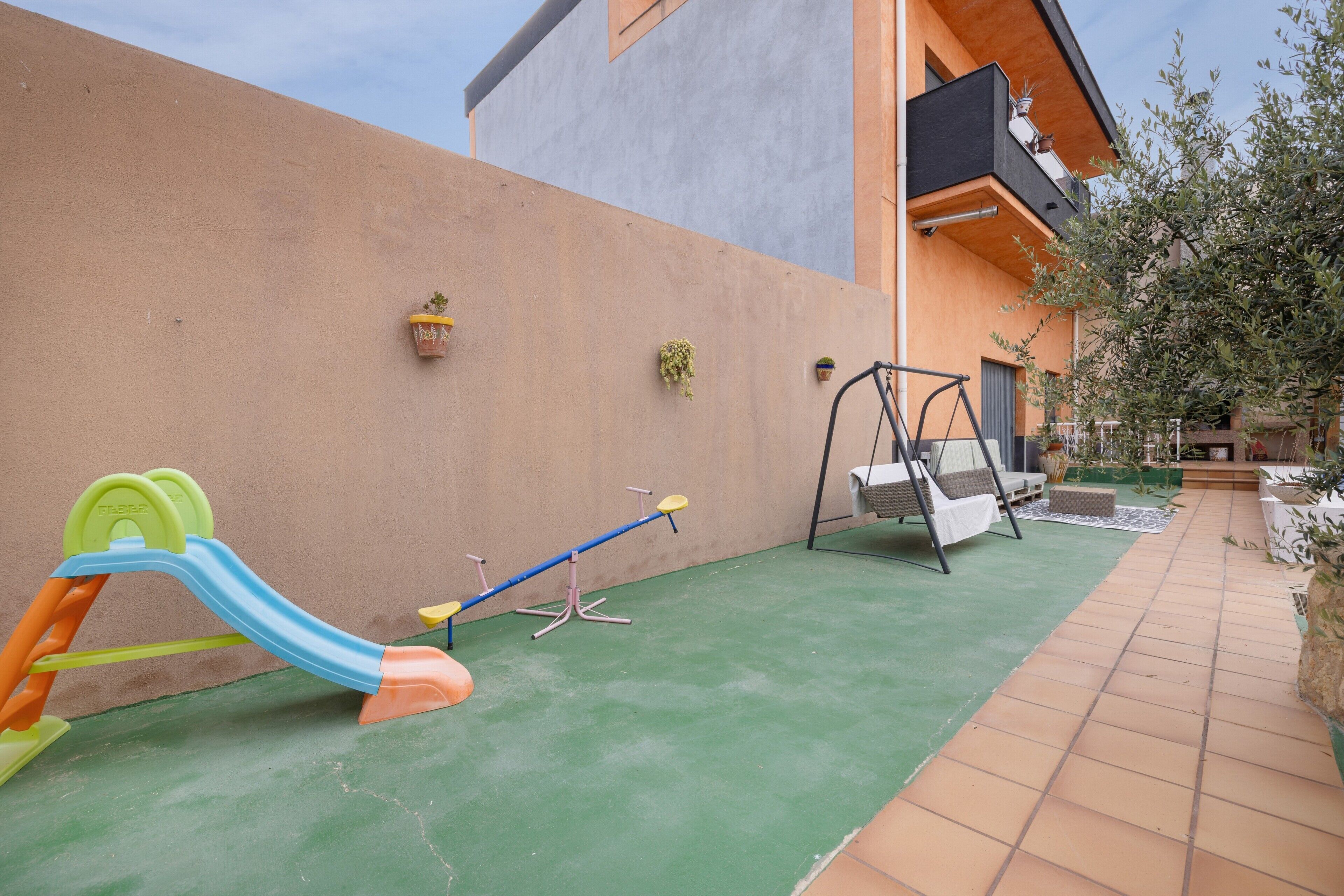 Un espace de jeux pour enfants avec un toboggan et une balançoire, un ensemble de balançoires et un pot de fleurs placé sur le mur extérieur d’un bâtiment.