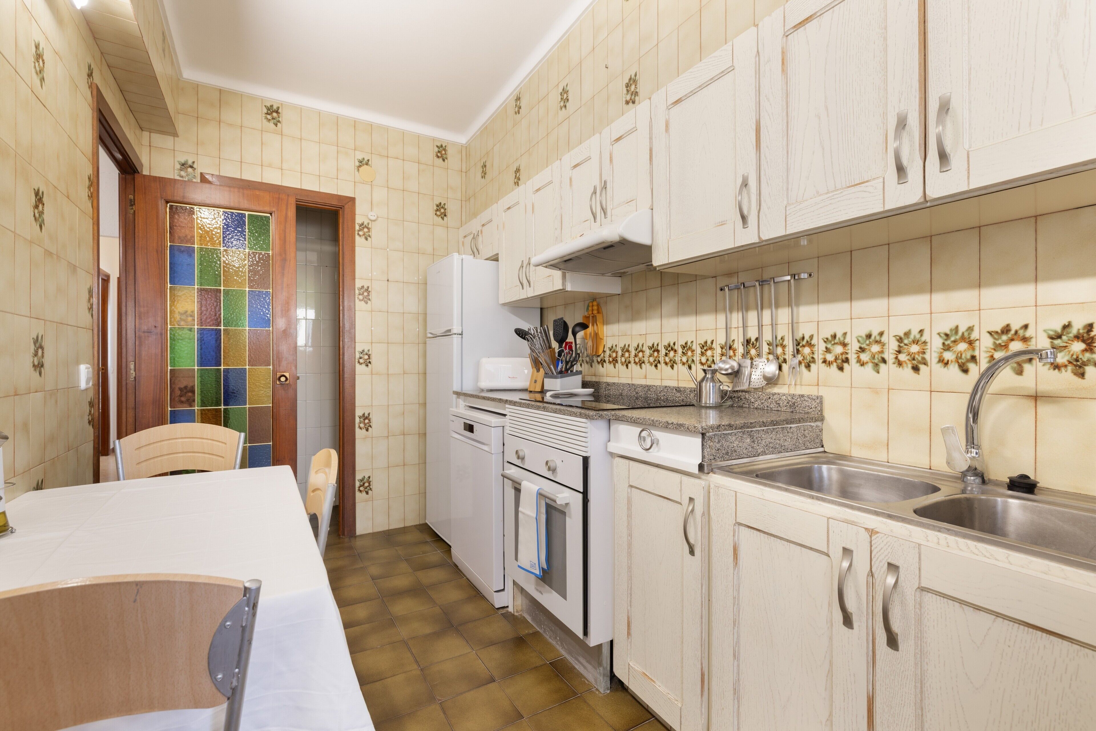 Una cocina con gabinetes blancos, salpicadero de azulejos y una puerta de madera con un panel de vidrio colorido.