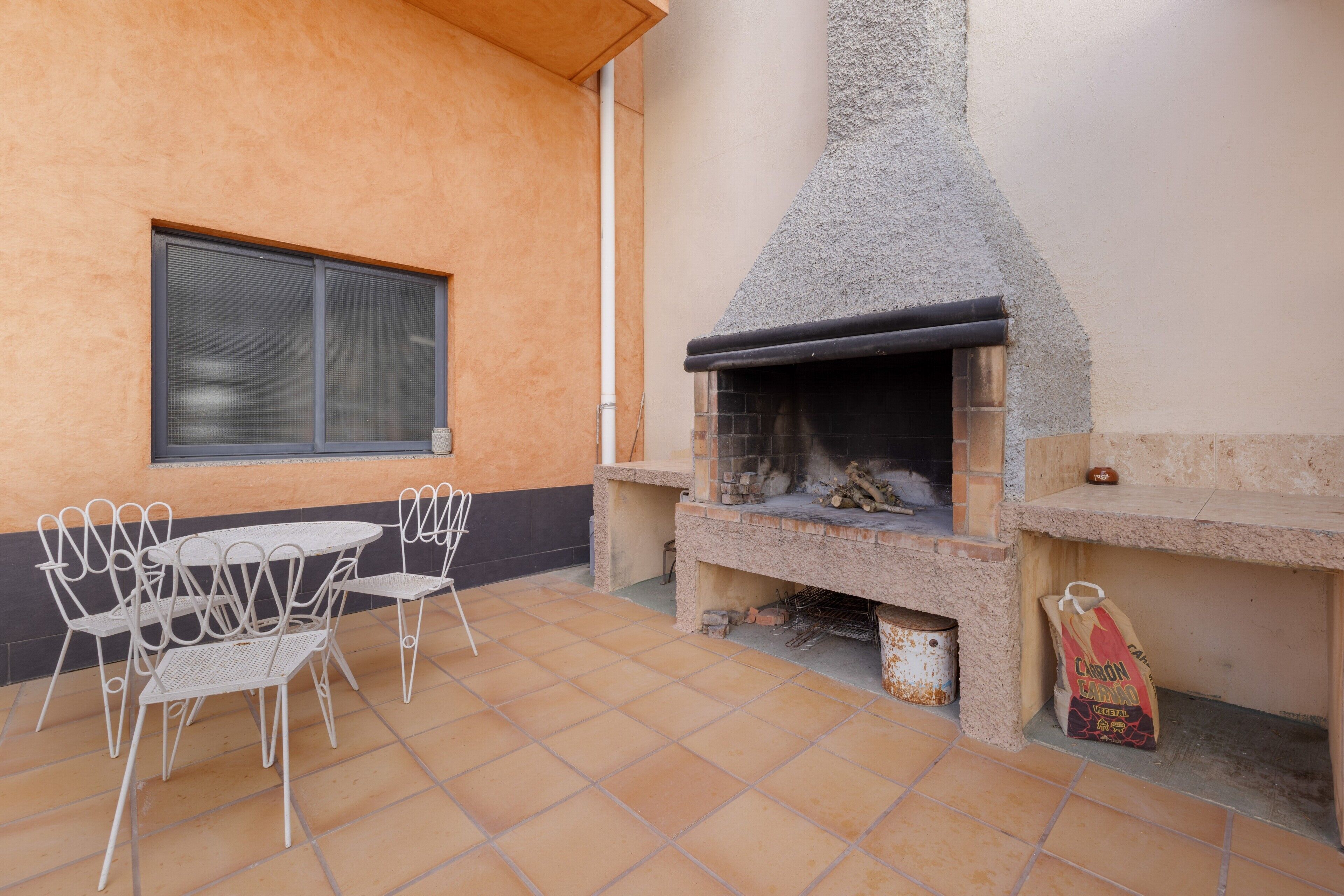 Eine Terrasse im Freien mit einem Stein-Barbecue, weißen Metallstühlen und einem Fenster.