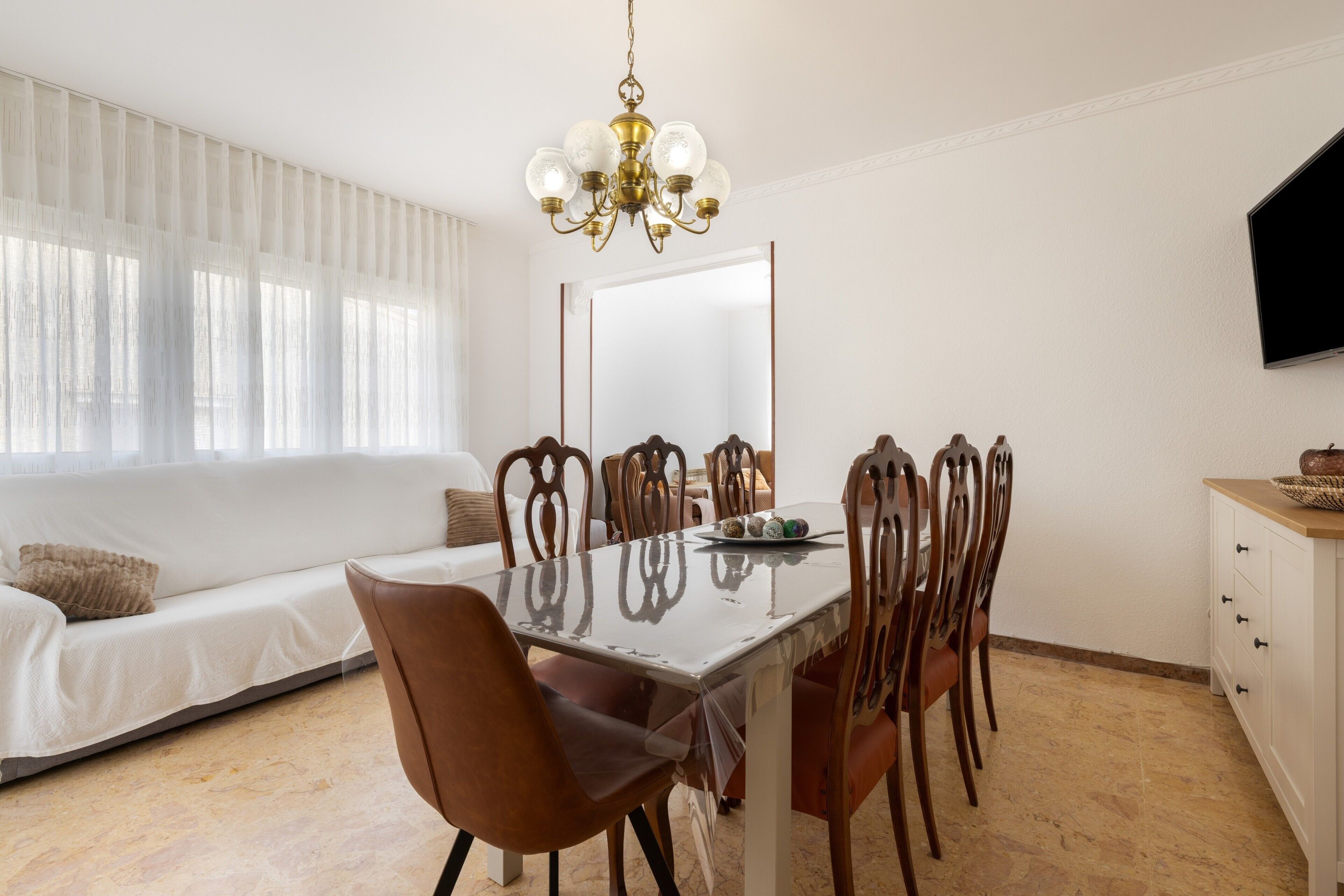 Uma sala de jantar com uma mesa, cadeiras, um lustre, um sofá e uma televisão.