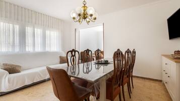 Uma sala de jantar com uma mesa, cadeiras, um lustre, um sofá e uma televisão.