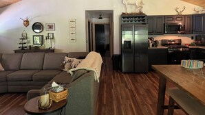 A cozy living room with a wooden ceiling, a gray sofa, a dining table, and a kitchen with dark cabinets and stainless steel appliances. - Welcome to 'Legacy Lodge',' your ultimate Centre County PA cabin getaway! (Munson)