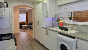 Una cocina compacta con lavadora integrada, un comedor con una mesa pequeña y sillas, y una sala de estar con sofá y ventana.