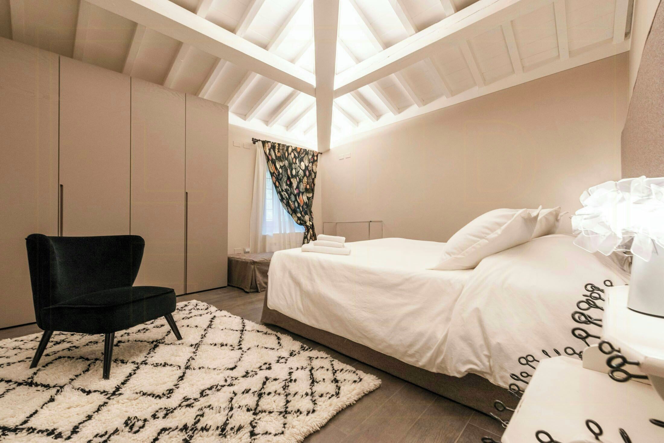 Une chambre à coucher comprenant un lit, une chaise, une table de chevet, une fenêtre avec des rideaux et un puits de lumière.