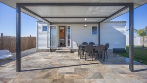 A covered patio with stone flooring, a table with chairs, and a white building with a sliding door. - Bungalow 'Tranquilo Cerca De Roche' with Private Pool, Wi-Fi and Air Conditioning (Conil de la Frontera)