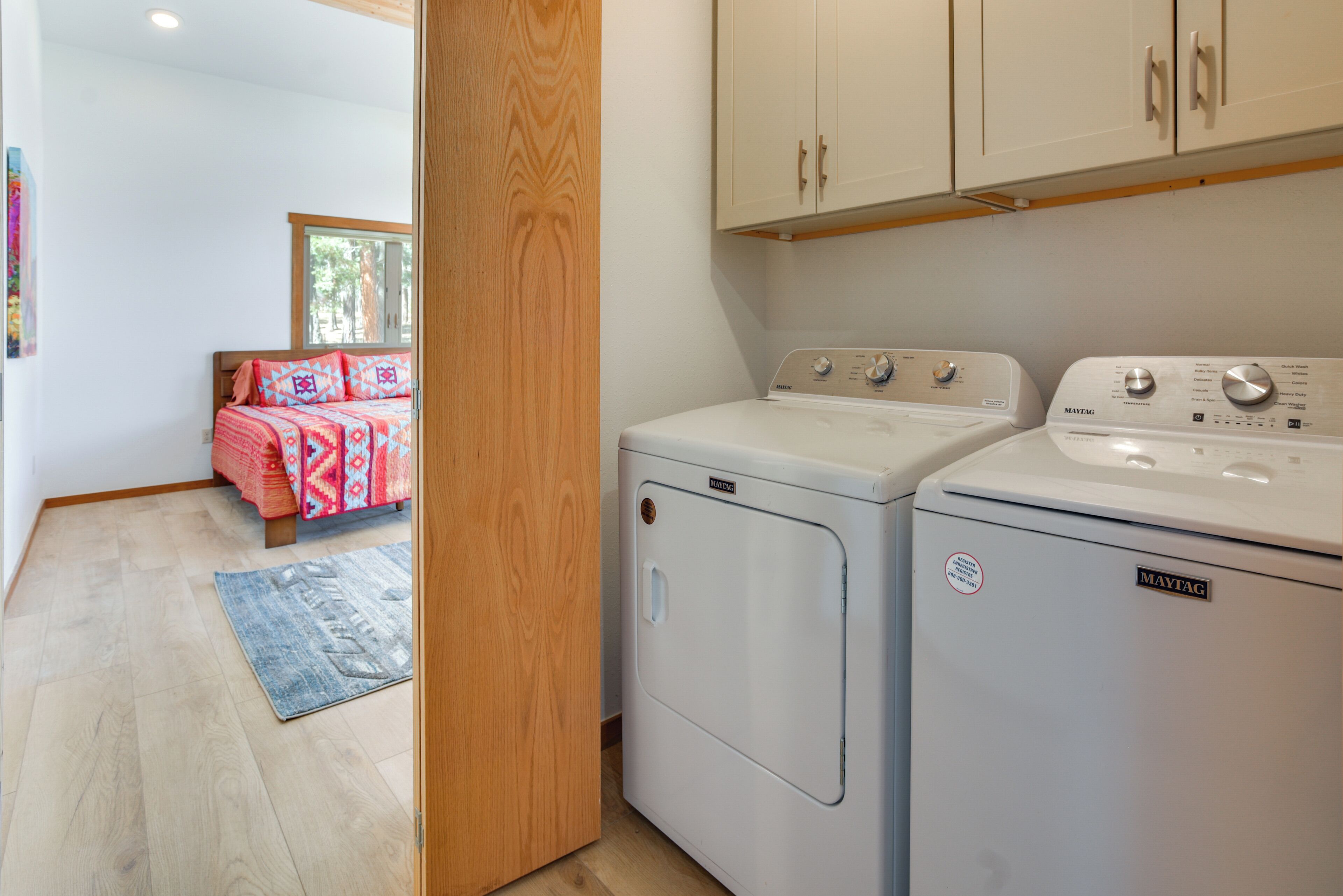 A laundry room with a washing machine and dryer, wooden flooring, and a bed with patterned bedding in the adjacent room.