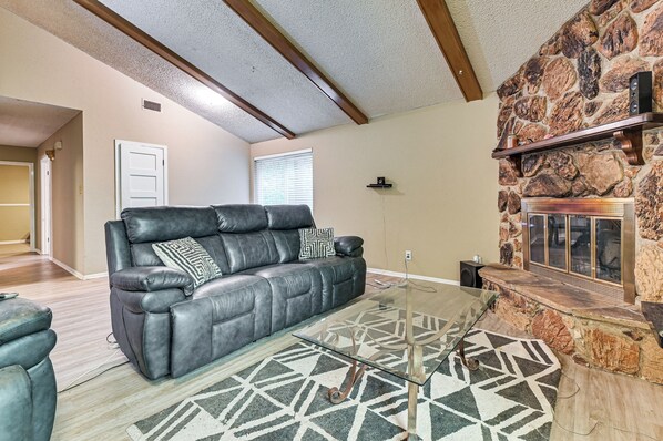 A living room with a leather sofa, a glass coffee table, a patterned rug, a stone fireplace, and a wooden ceiling beam. - 2 Mi to Denton Square! Spacious Home w/ Backyard (Denton)