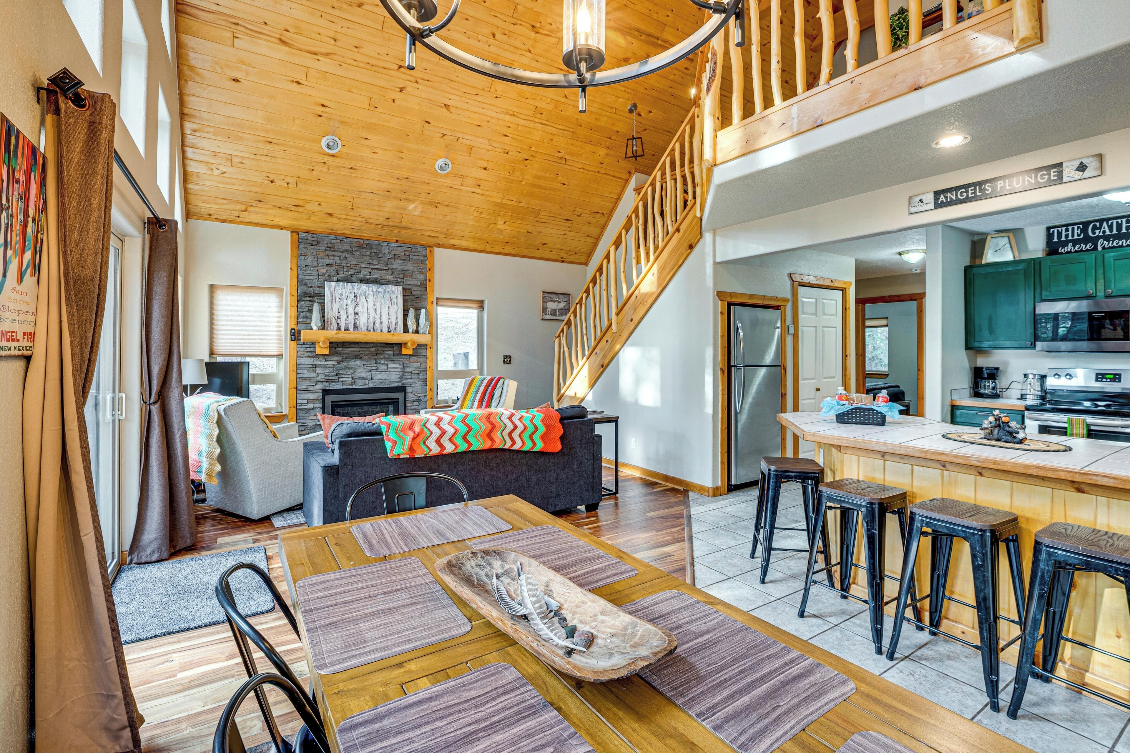 A spacious living area with a wooden ceiling, a stone fireplace, a dining table with stools, and a kitchen area with a bar.