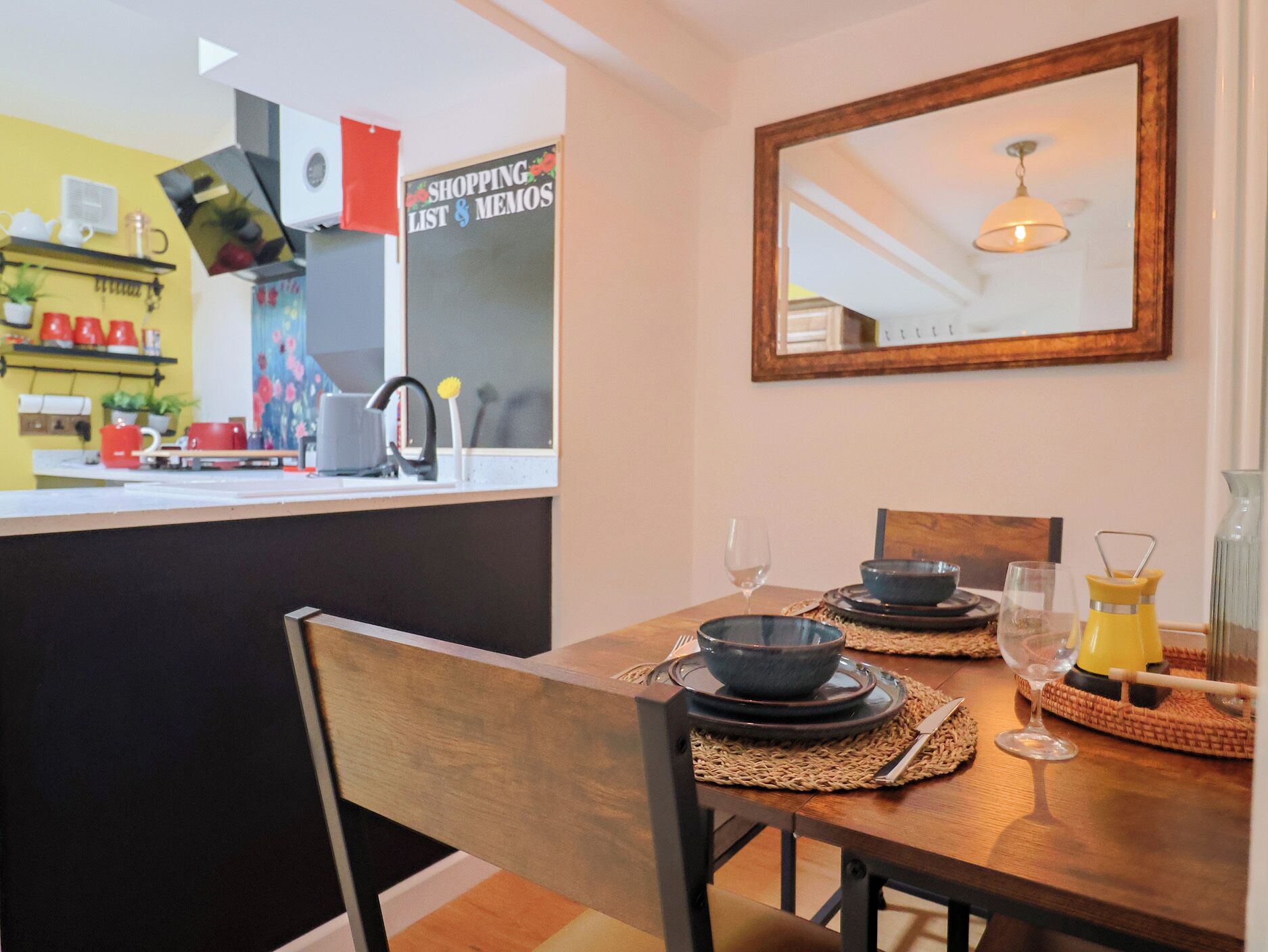 A dining area with a table set for two, a mirror on the wall, and a kitchen in the background.