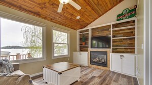A living room with a fireplace, wooden ceiling, and large windows overlooking a body of water. - Dock, Deck & Sunsets! Little Bungalow on Lake Mary (Twin Lakes)