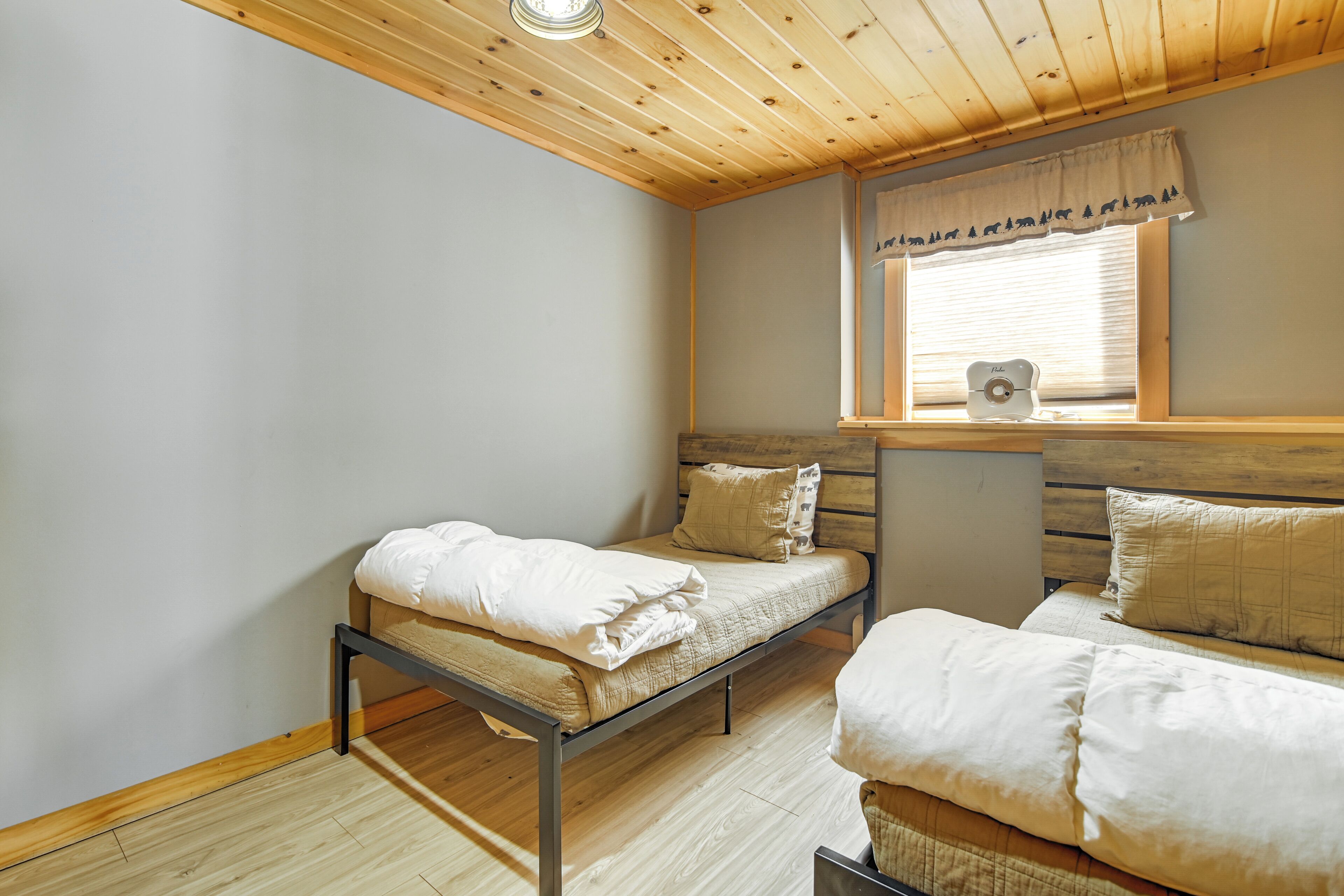 A room with two beds, wooden ceiling, a window with blinds, and a wall-mounted heater.
