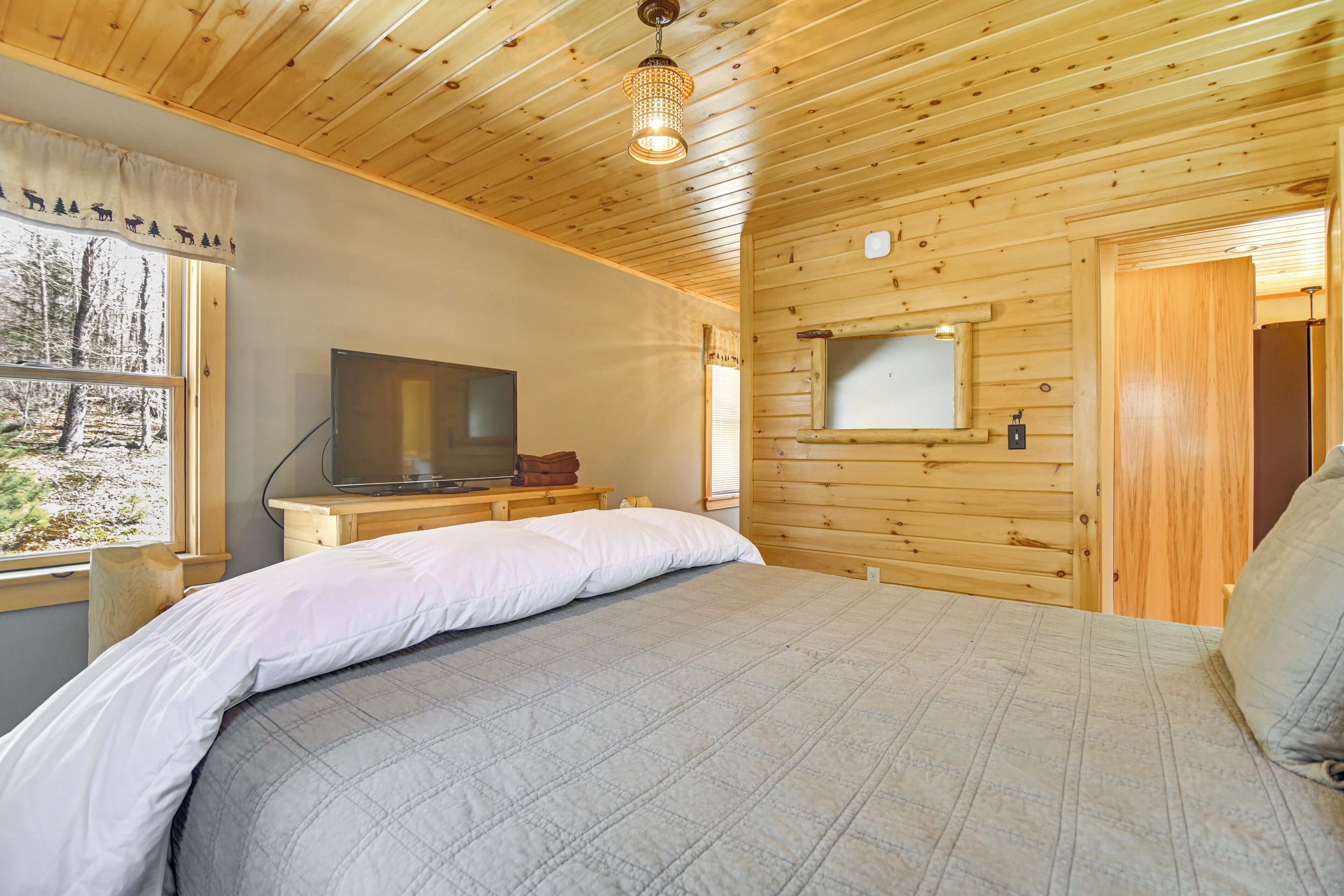 A wooden cabin bedroom with a bed, a television, and a window overlooking a forest.