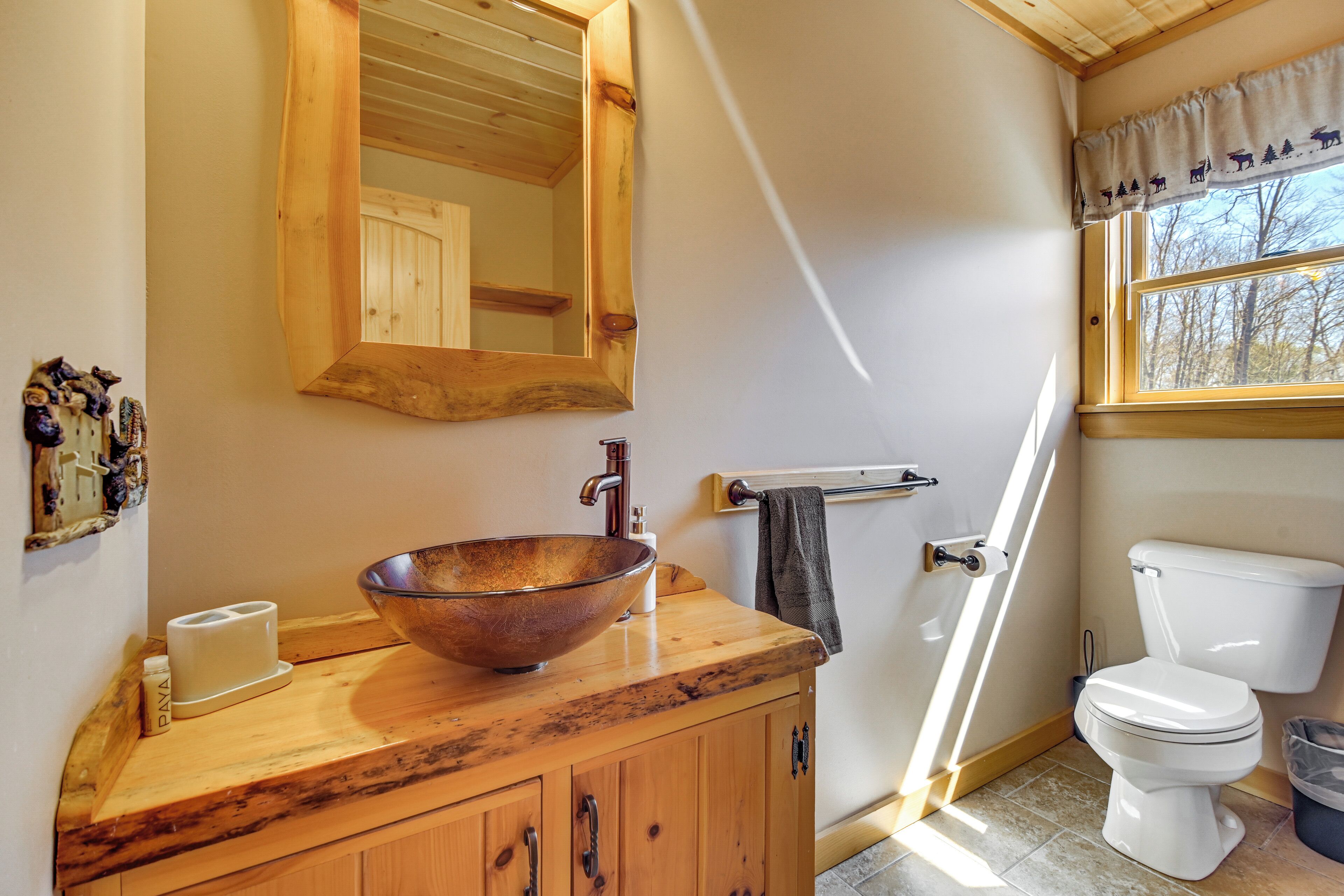 A bathroom with a wooden vanity, a large mirror, a vessel sink, a toilet, and a window with a view of trees.