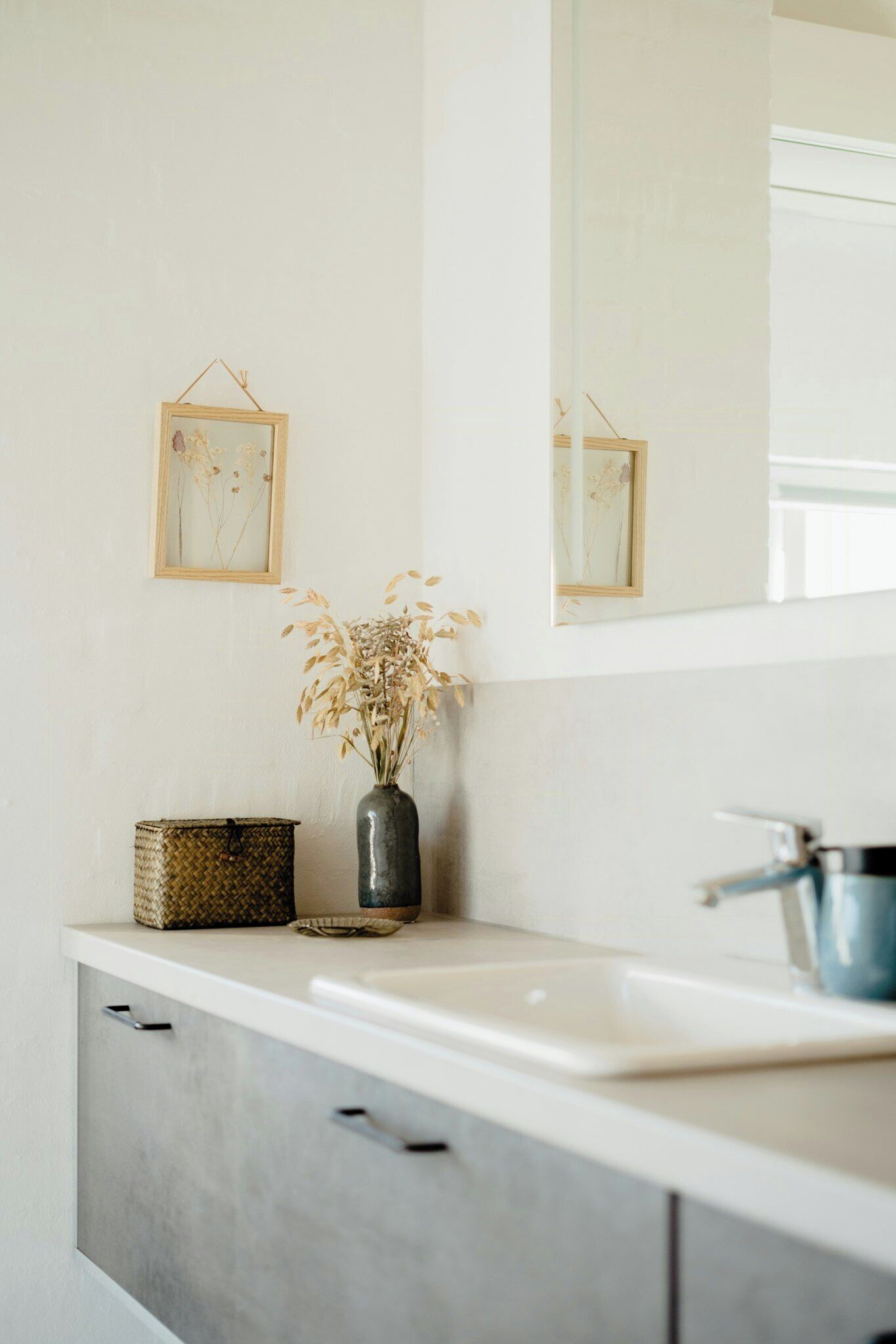 Une salle de bain avec un lavabo blanc, un miroir, un vase contenant des fleurs séchées et un panier en osier.