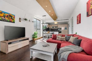 A modern living room with a flat-screen TV, a red sofa, a coffee table, and a kitchen in the background.