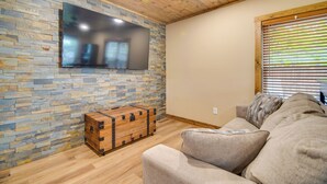 Un salon avec un mur en pierre, un plafond en bois, un téléviseur à écran plat, une commode en bois et un canapé gris.