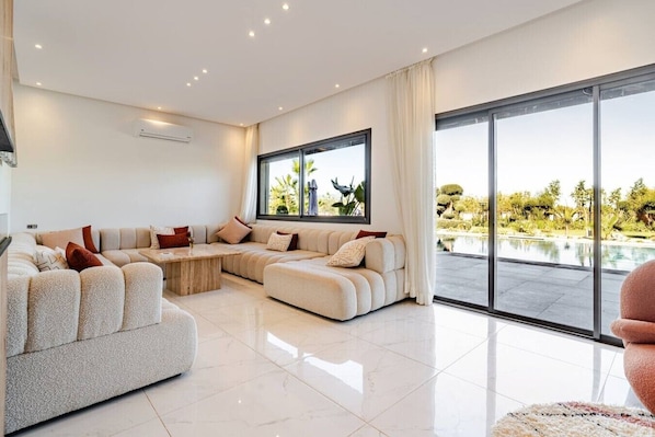 A modern living room with a sectional sofa, a coffee table, and a large window with a view of a pool and greenery. - Pool • Retreat at Villa Taroub • 4 Suits • Gym (Marrakesh)