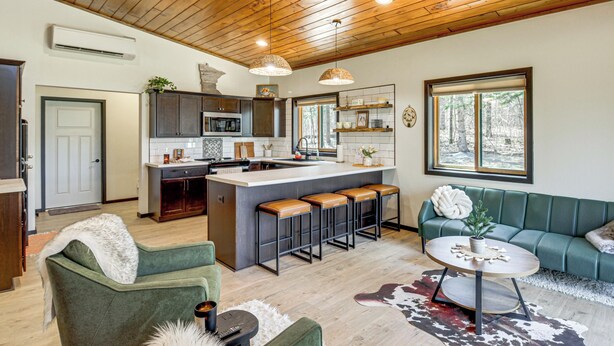 A modern kitchen with a central island, bar stools, and a green sofa in a spacious living area with wooden flooring and a high ceiling. - Long Lake Cabin w/ Dock: Fish & Hike! (Akeley)