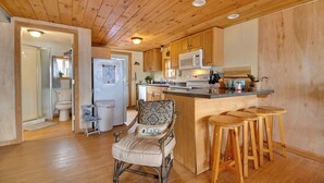 A wooden interior with a kitchen, a dining area with stools, and a chair with a pillow.