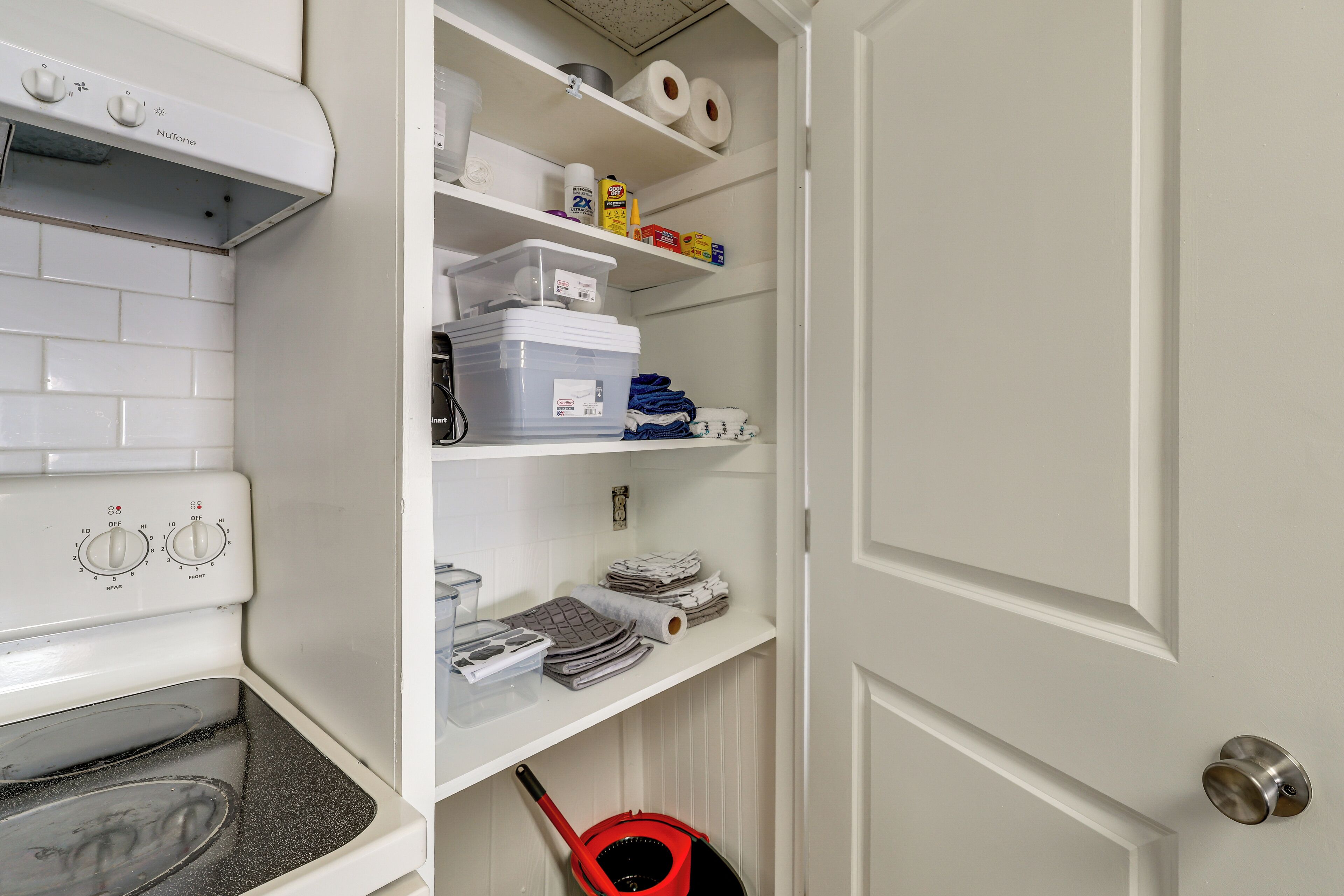 A kitchen with a built-in oven, a white door with a silver handle, and shelves with various items including paper towels and cleaning supplies.