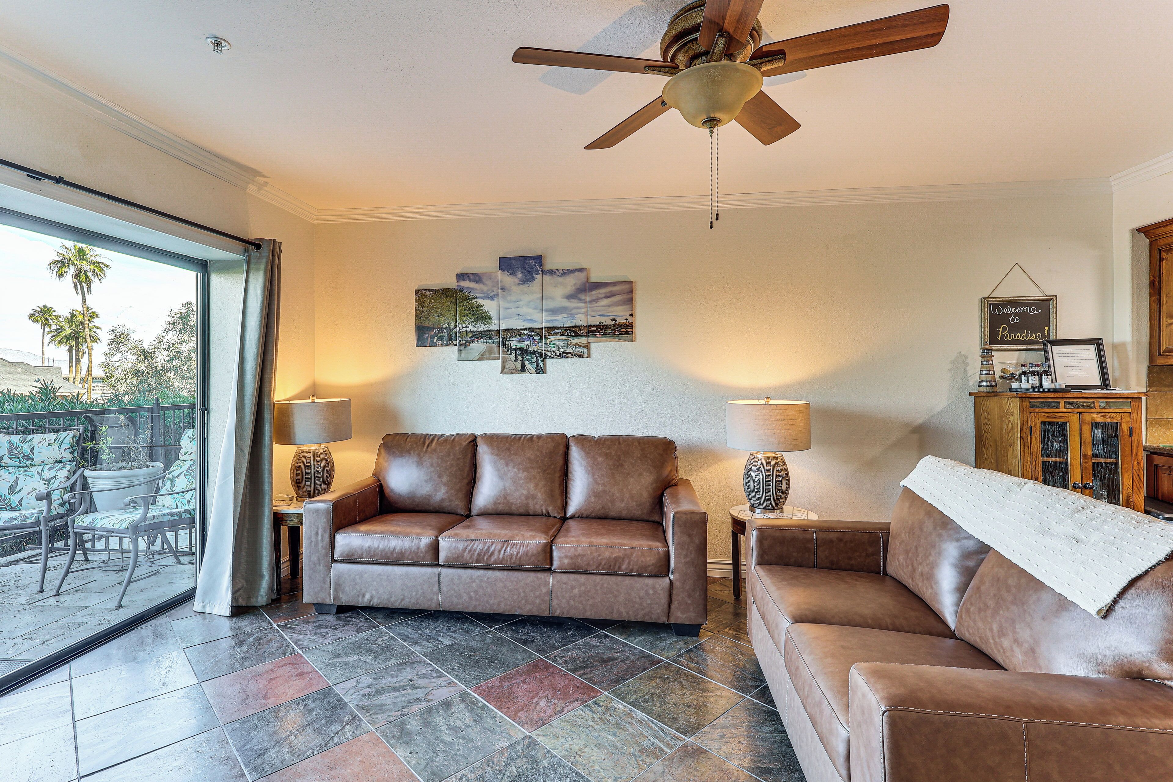 A living room with a brown leather sofa, two side tables with lamps, a ceiling fan, a wall-mounted painting, and a view through a glass door to an outdoor patio.