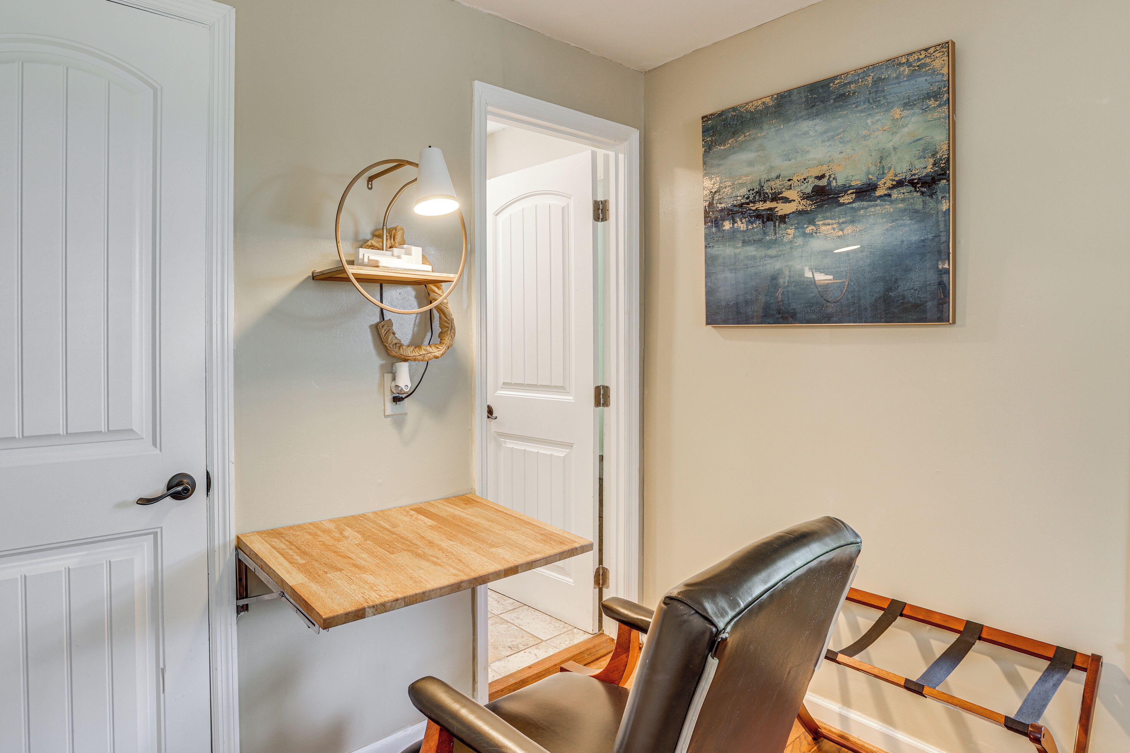 A room with a wooden desk, a chair, a wall-mounted lamp, and a painting.