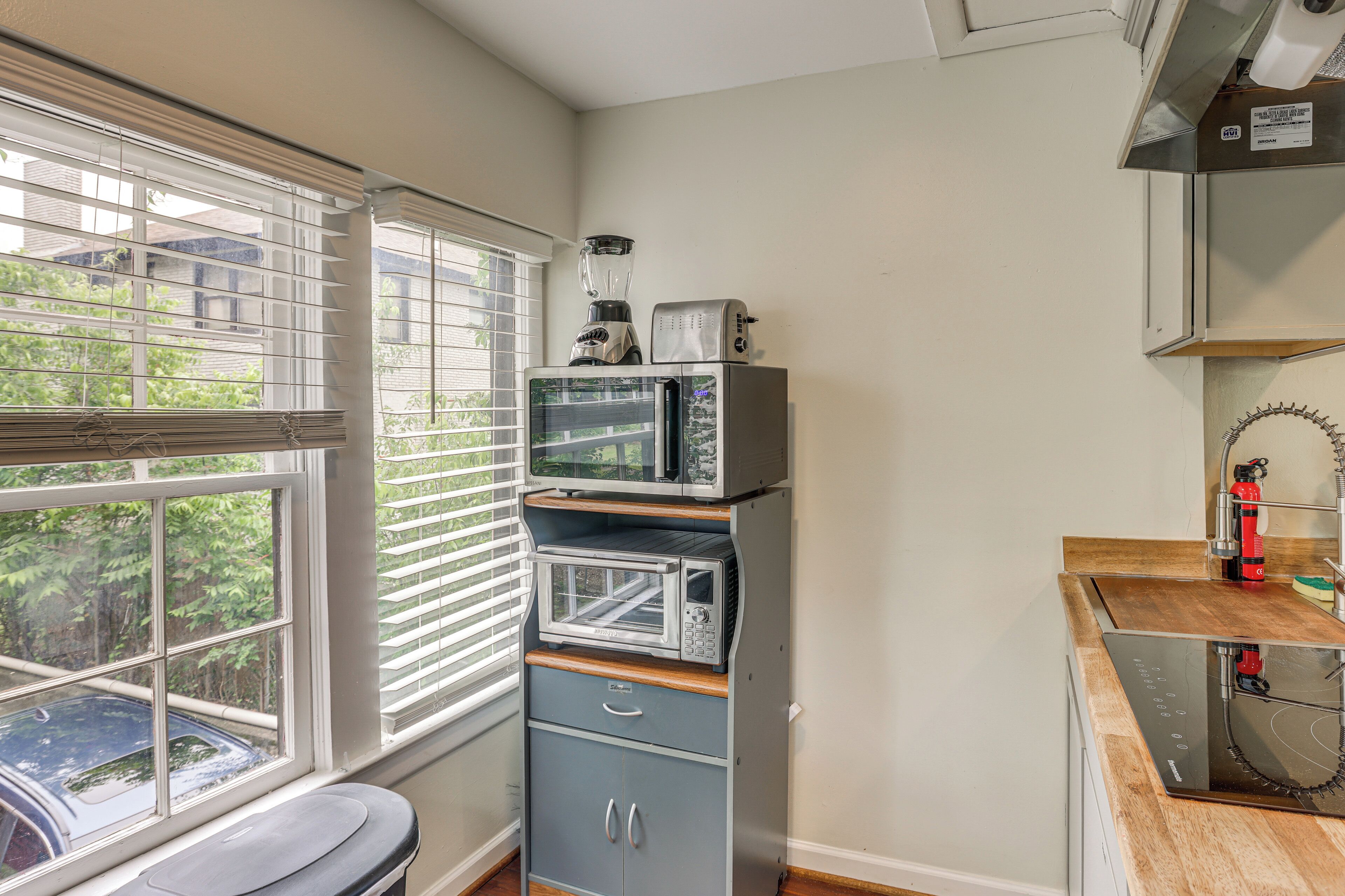 A kitchen with a microwave, toaster, and blender on a cabinet, a window with blinds, and a view of a car outside.