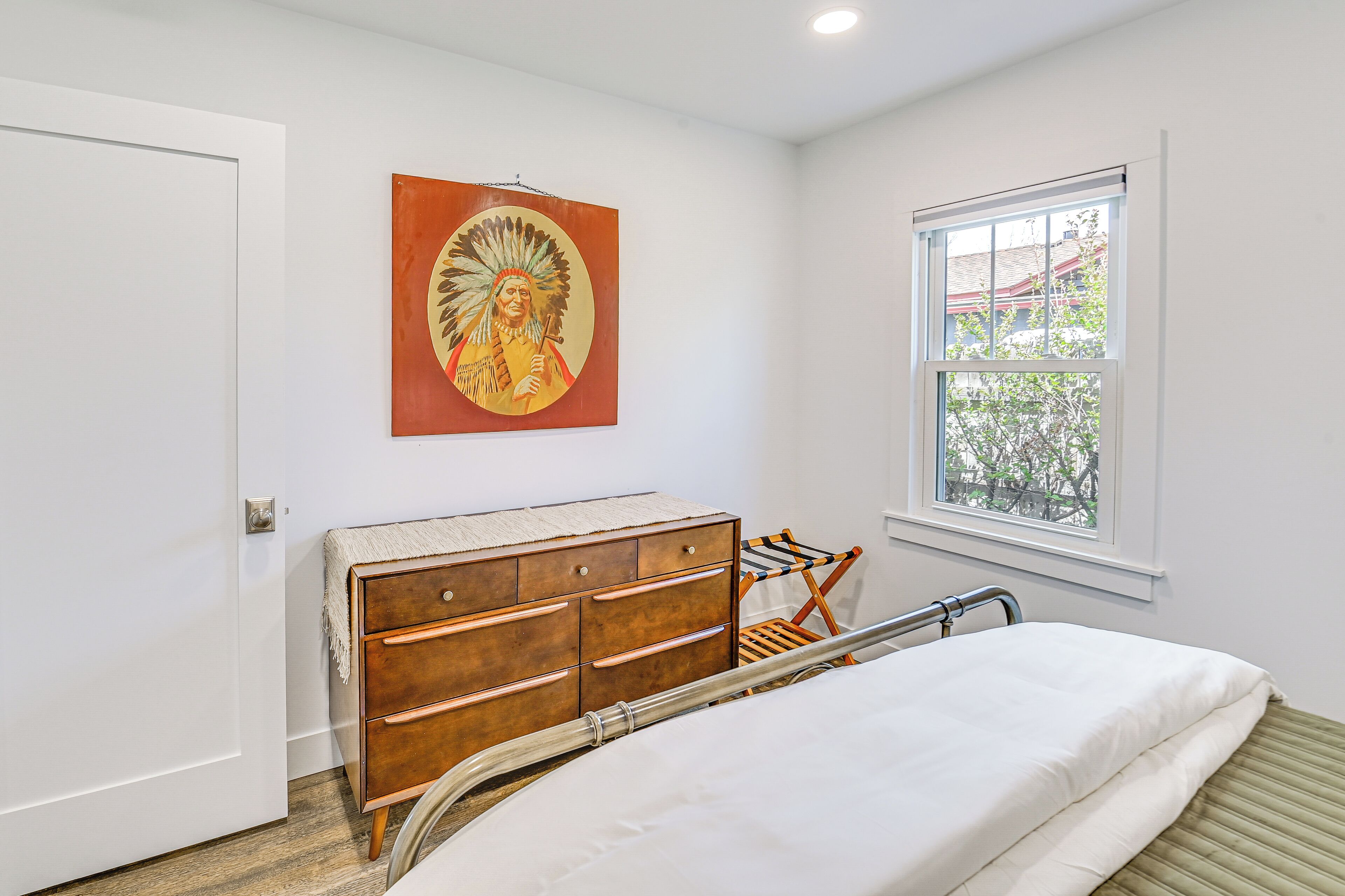 A bedroom with a bed, wooden dresser, a window with a view, and a wall painting.