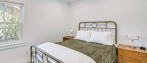 A bedroom with a metal bed frame, a green bedspread, white pillows, a wooden nightstand with a lamp, and a window with a view of trees.