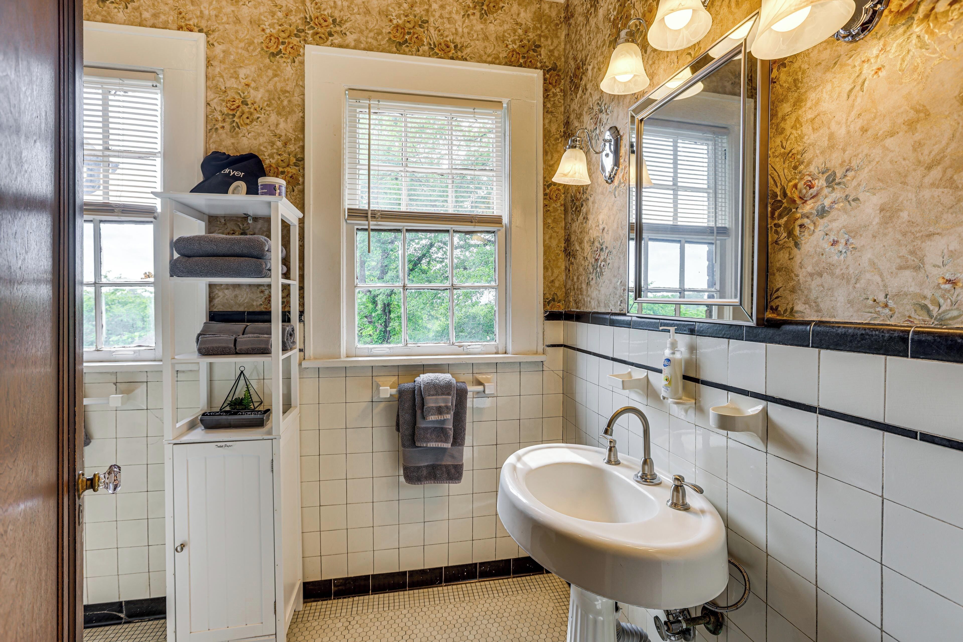 A bathroom with a white sink, a mirror, a window with blinds, and a shelf with towels.