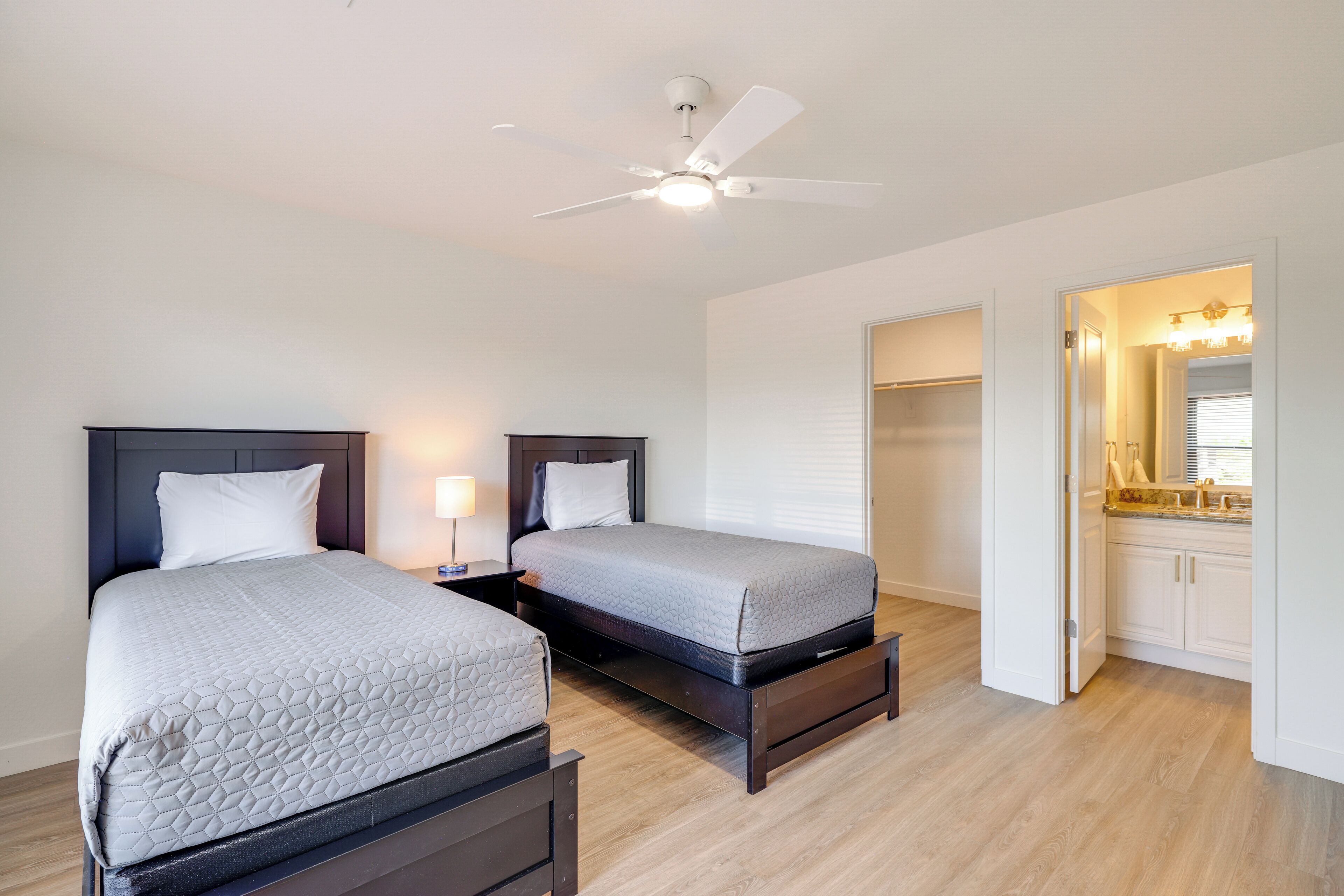 A bedroom with two beds, a ceiling fan, a nightstand, and a bathroom with a vanity and mirror.