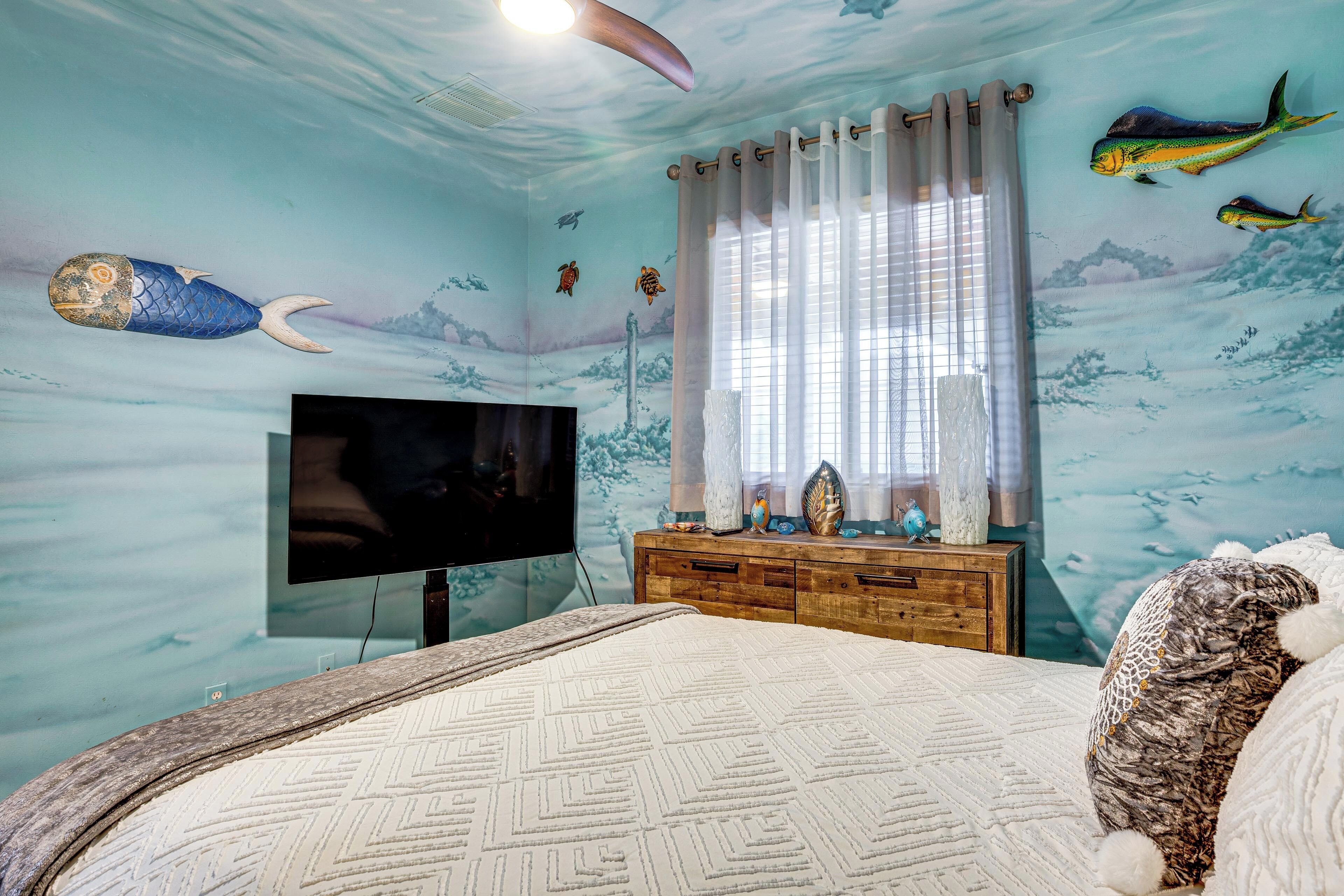 A bedroom with a bed, a TV, a wooden dresser, and a window with curtains.