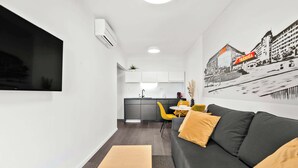 Un salon moderne avec un canapé, un téléviseur fixé au mur et une cuisine en arrière-plan.