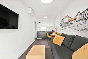 Ein modernes Wohnzimmer mit einer Couch, einem an der Wand befestigten Fernseher und einer Küche im Hintergrund.