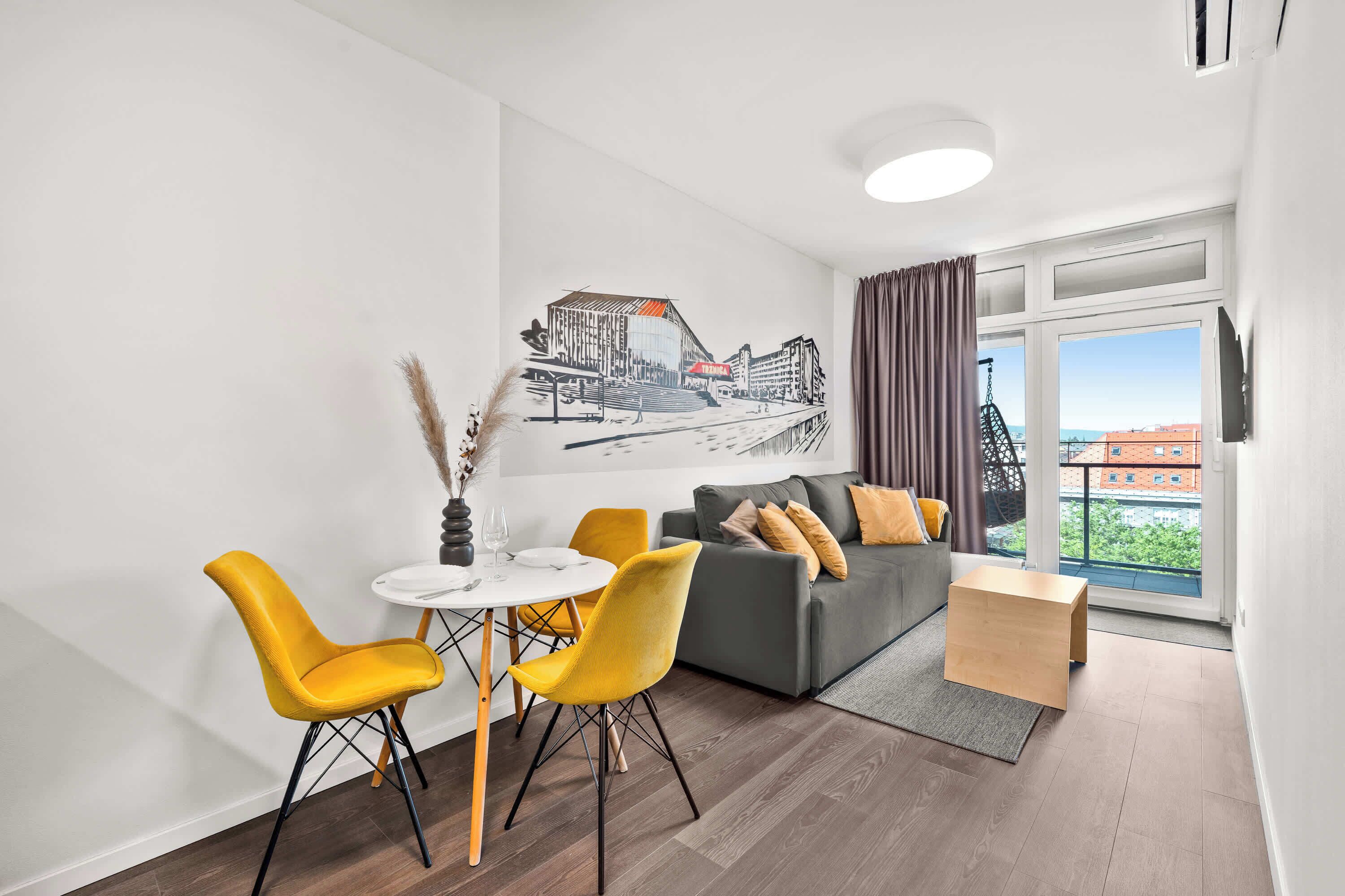 Un salon moderne comprenant un coin repas, un canapé avec des coussins, une petite table et une grande fenêtre donnant sur un beau paysage.