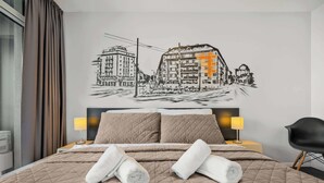 Une chambre d’hôtel comprenant un lit, deux oreillers, une tête de lit, une table de chevet, une lampe, une chaise et une œuvre d’art murale représentant des bâtiments.