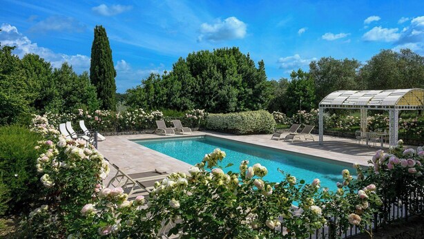 Piscina con sillones de descanso y una pérgola, rodeada de vegetación exuberante y plantas florecientes.