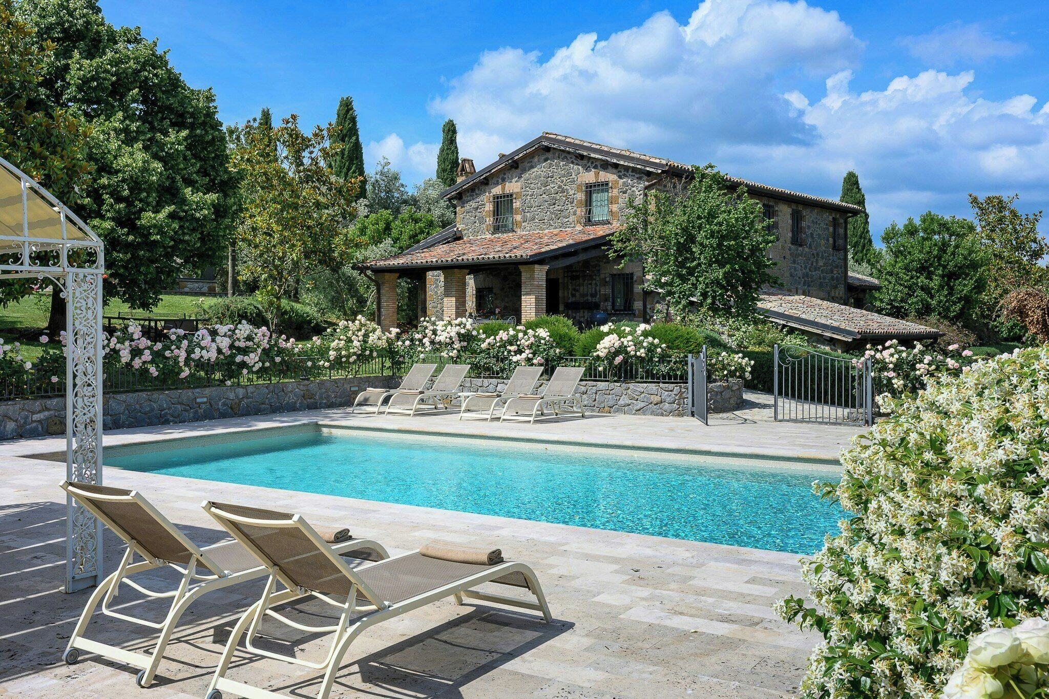 A stone building with a swimming pool, surrounded by lush greenery and white flowers.