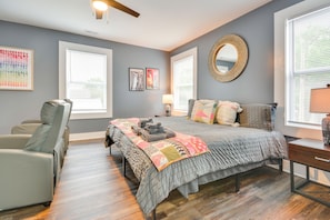 A bedroom with a bed, two windows, a chair, a bedside table, and a mirror.
