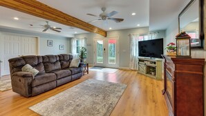 A living room with a brown sofa, wooden beams, ceiling fans, a TV, and a wooden cabinet. - Fire Pit & Forest Views: Lakes Region Retreat (Holderness)