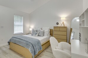 Une chambre à coucher comprenant un lit, une table de chevet, une lampe, une chaise, une commode et une fenêtre avec des stores.
