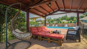 Ein überdachter Außenbereich mit Hängestuhl, einer roten gepolsterten Couch und einem Pool im Hintergrund.
