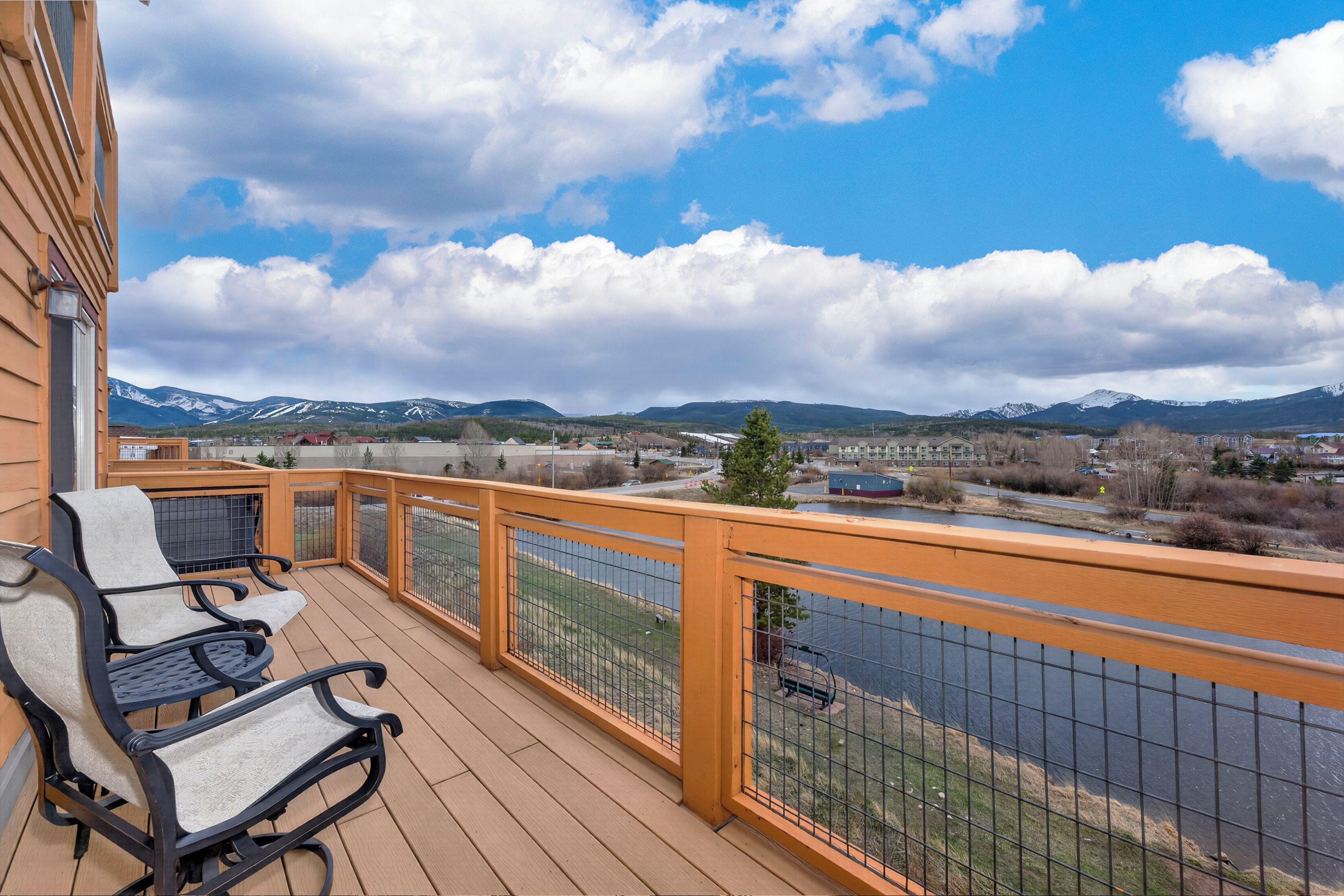 A wooden deck with outdoor furniture, a metal railing, and a view of mountains and a river.