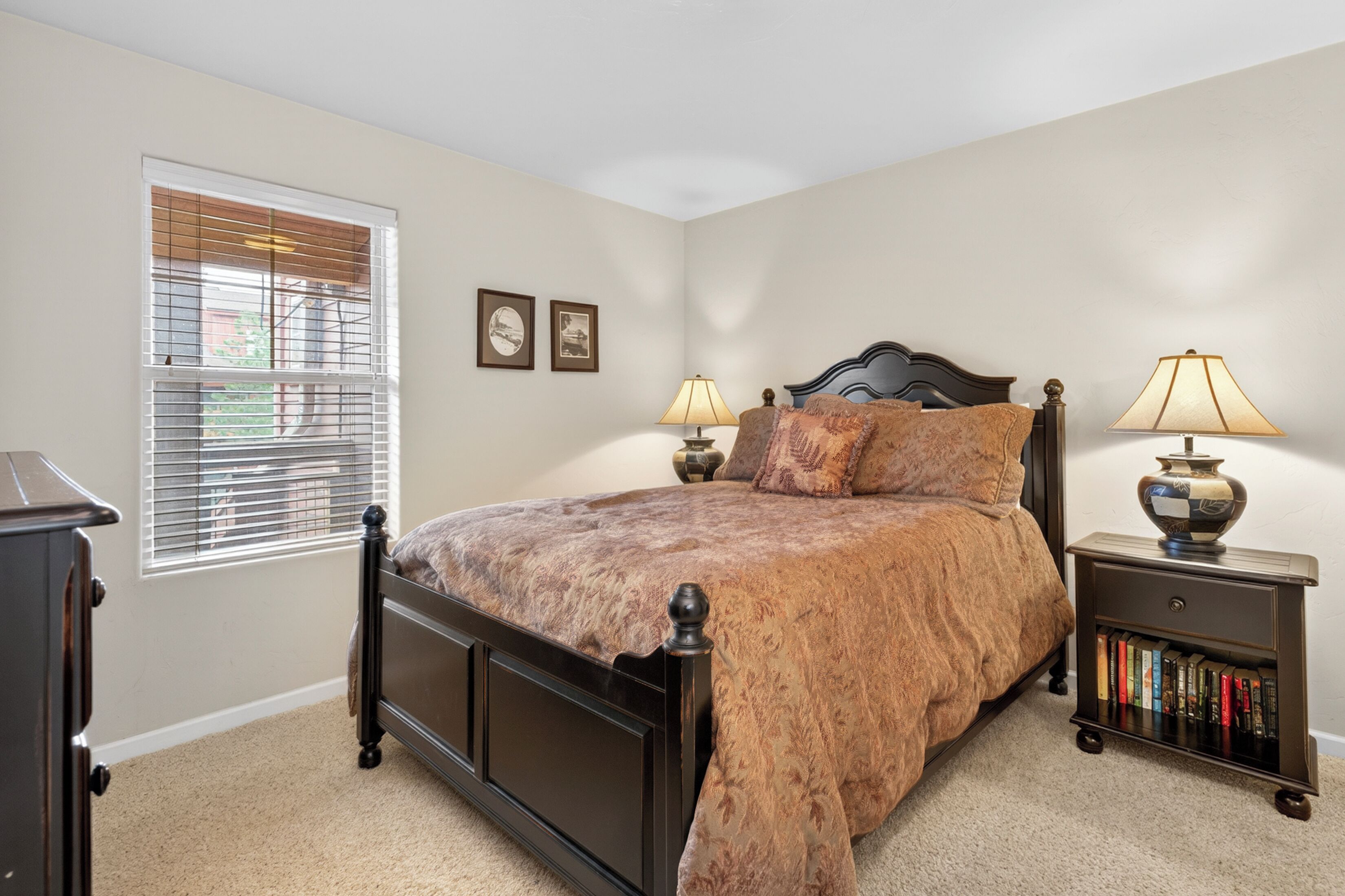 Quarto com cama, duas luminárias de cabeceira, mesa de cabeceira com livros e janela com persianas.